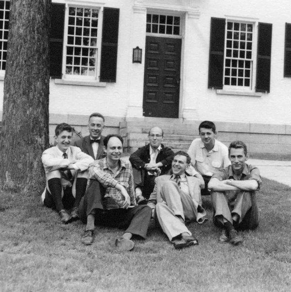 black and white photo of seven smiling men sitting on a lawn in-front of a tree and a white school building with many windows