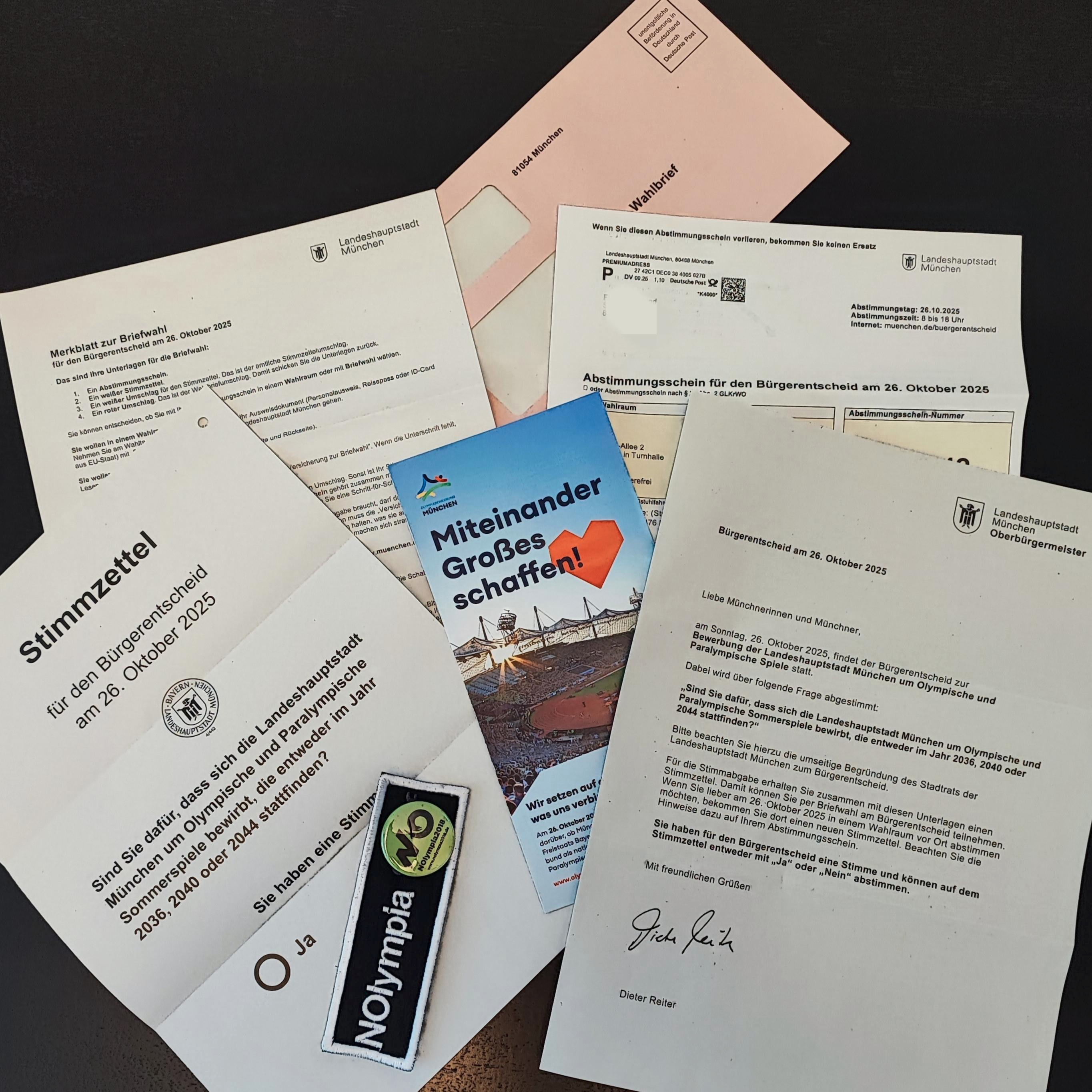 Foto des Inhalts der Abstimmungsunterlagen. Stimmzettel, Merkblatt, Wahlschein, Wahlbriefkuvert, Pro-Flyer und Anschreiben OB.  Oben drauf gelegt: ein NOlympia Aufnäher.